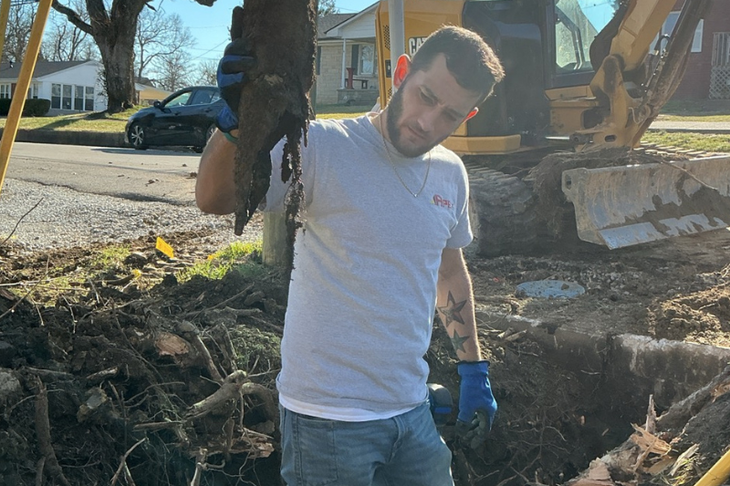 Technician Aaron holding up a broken pipe. He's standing in a trench overed in cut up roots.